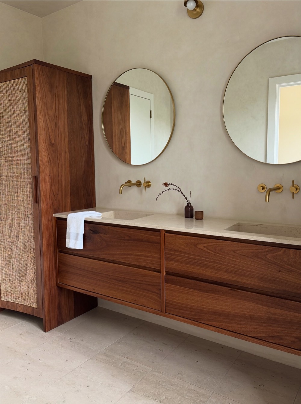 A floating warm wood double vanity with a pale stone-look top, gold-toned wall faucets and two round mirrors.