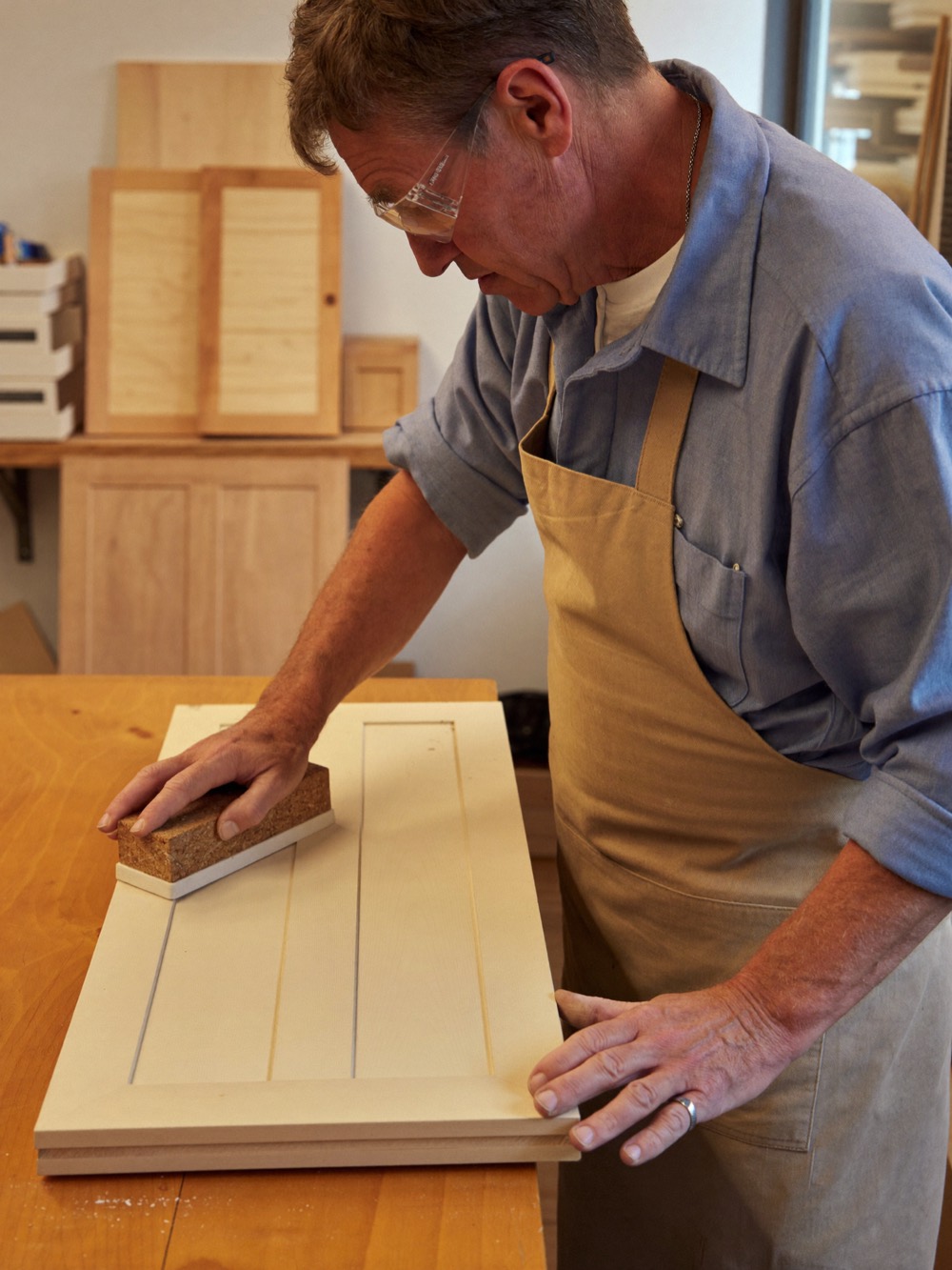 A woodworker in safety glasses and a canvas apron hand-sands a painted shaker cabinet door on a workbench.