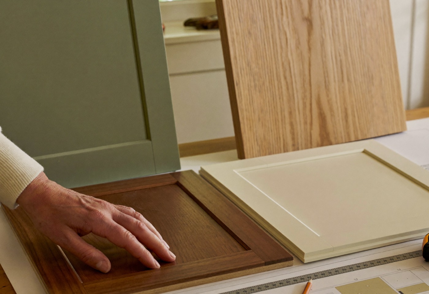 A hand rests on a dark wood cabinet door sample beside ivory, sage and pale wood samples on a table spread with kitchen plans.