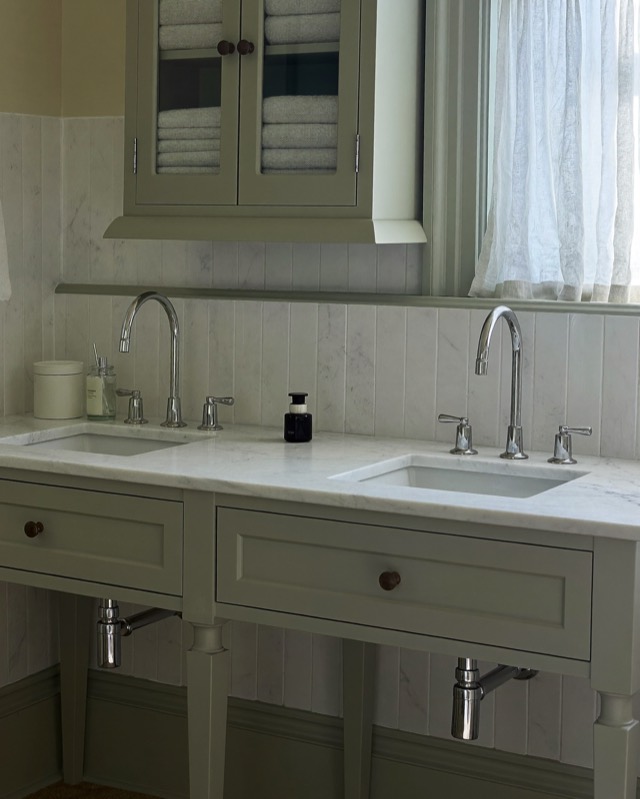 A pale green-grey painted double vanity on legs with a white veined top, silver-toned faucets and a glass-front towel cabinet above.
