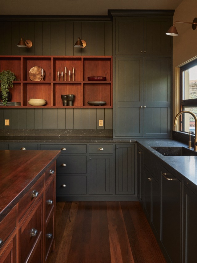 Deep green-black painted cabinetry with dark stone-look counters, a warm wood open shelf and a wood island in the foreground.