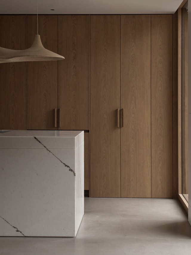 Floor-to-ceiling pale wood flat-panel cabinetry with long wooden pulls behind a veined stone-look island.