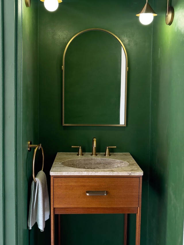 A small warm wood vanity with one drawer and a carved stone-look basin against deep green walls, under an arched gold-toned mirror.