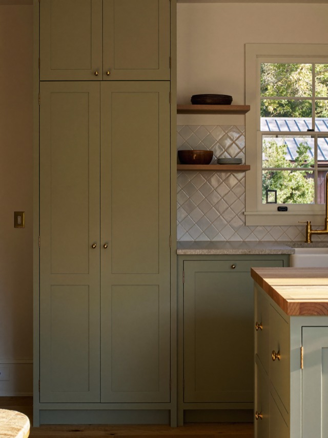 Sage green painted shaker cabinetry with gold-toned knobs, a wood-topped island and open wood shelves by a window.
