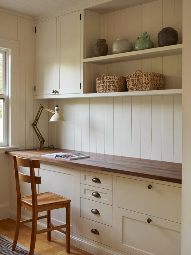 An ivory painted built-in study wall with a warm wood desktop, a bank of drawers with cup pulls and open shelves holding baskets and vases.