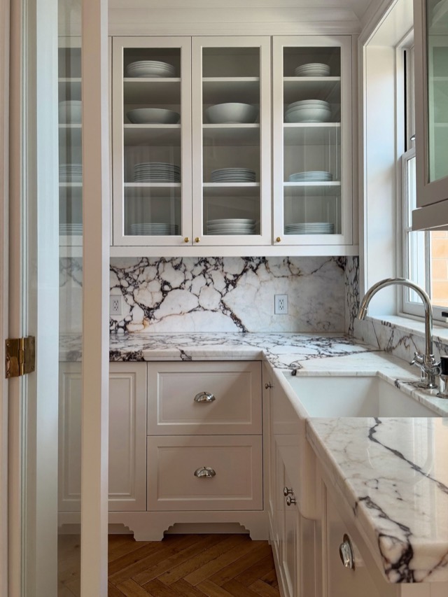White painted inset cabinetry with glass-front uppers, silver-toned cup pulls and boldly veined stone-look counters beside a window sink.