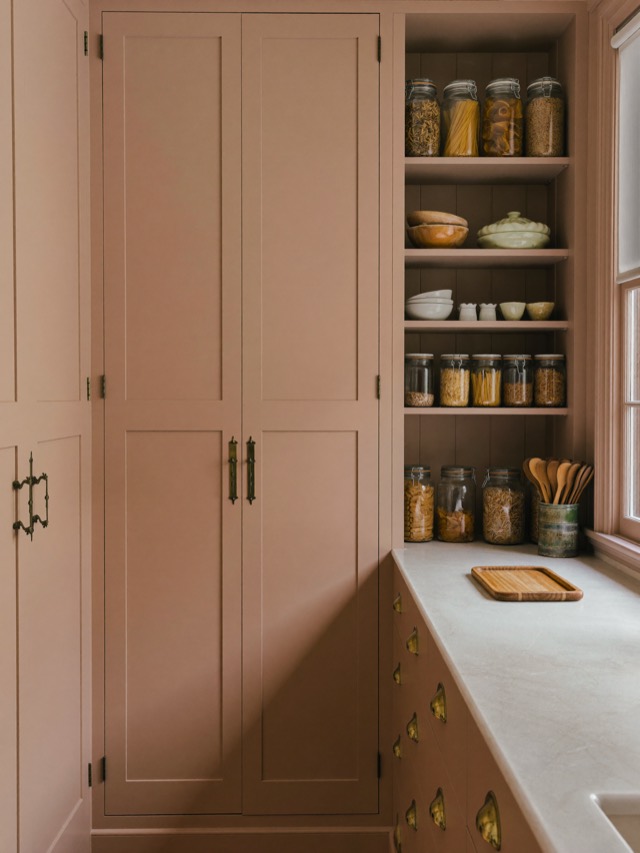 A walk-in pantry with tall painted shaker doors, open shelves of glass jars and drawers with gold-toned cup pulls under a pale stone-look counter.
