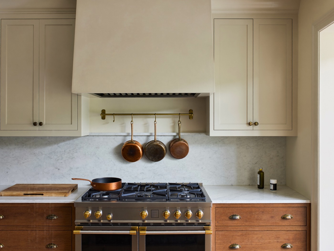 A cooking wall with an ivory range hood and upper cabinets, warm wood drawers with gold-toned cup pulls and a veined stone-look backsplash.