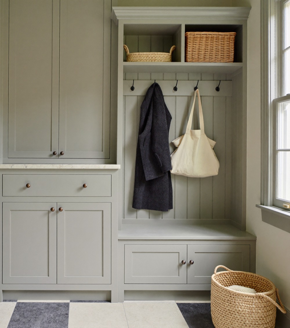 Sage grey painted mudroom cabinetry with a built-in bench, coat hooks and baskets beside a window.