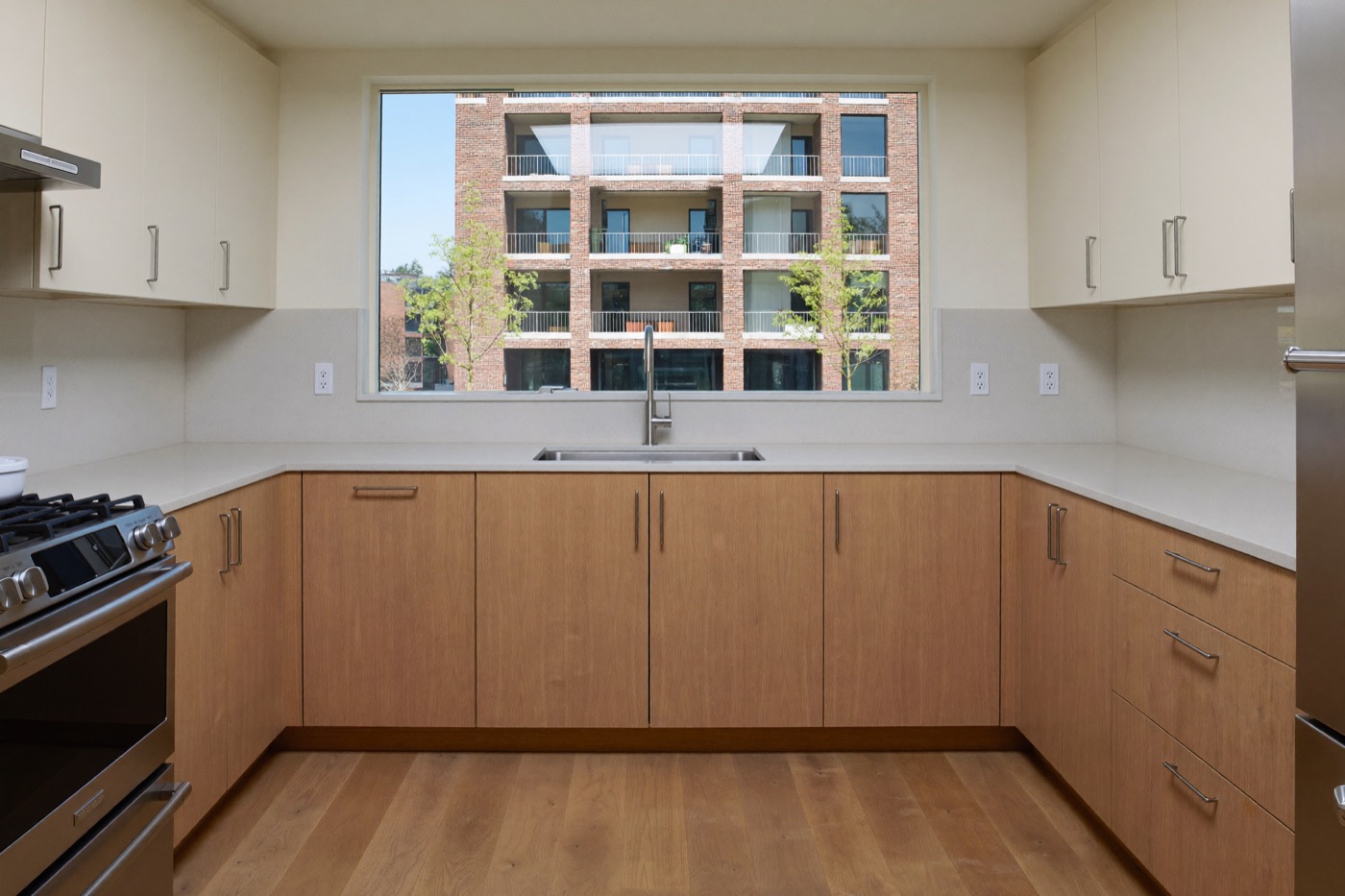 A newly finished apartment kitchen with pale wood lower cabinets and ivory uppers, and a window facing a brick apartment building.